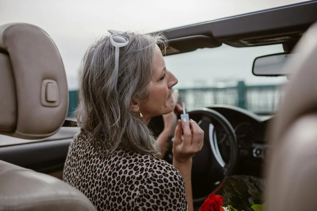 Side view of middle aged female with gray hair in stylish outfit applying lip gloss while driving modern cabriolet